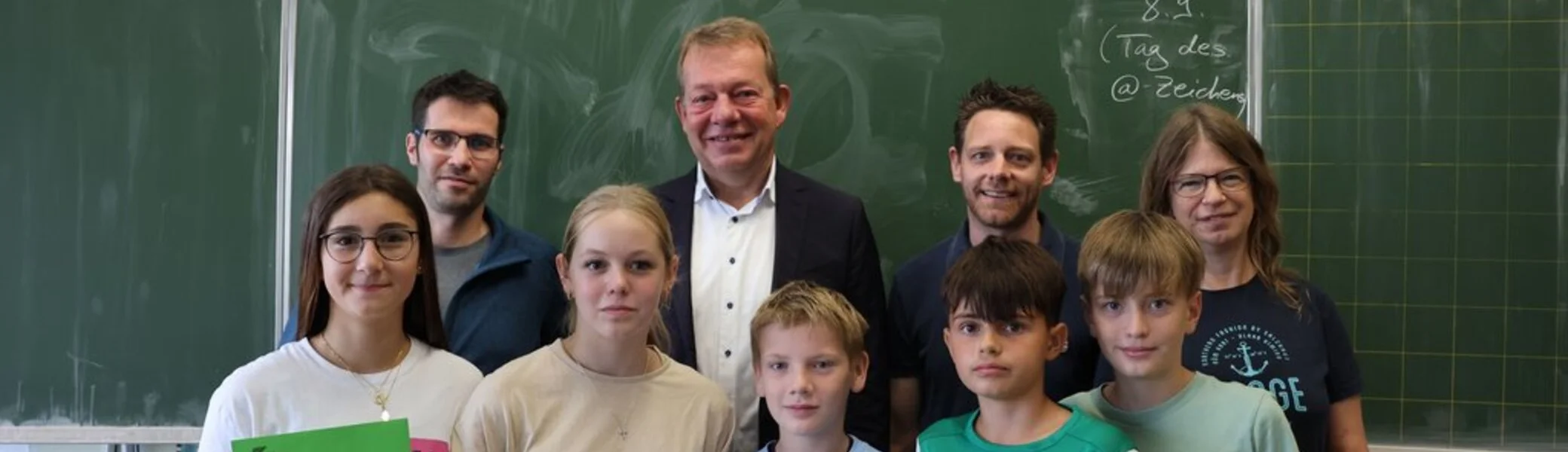 Eine Gruppe steht vor einer Tafel in einem Klassenraum, in der hinteren Reihe stehen Erwachsene in der vorderen Reihe 5 Schüler/innen. Die Schüler/innen halten ein Plakat mit der Aufschrift "Herzlichen Glückwunsch" und einen grünen Umschlag in der Hand.  | © Universitätsstadt Siegen