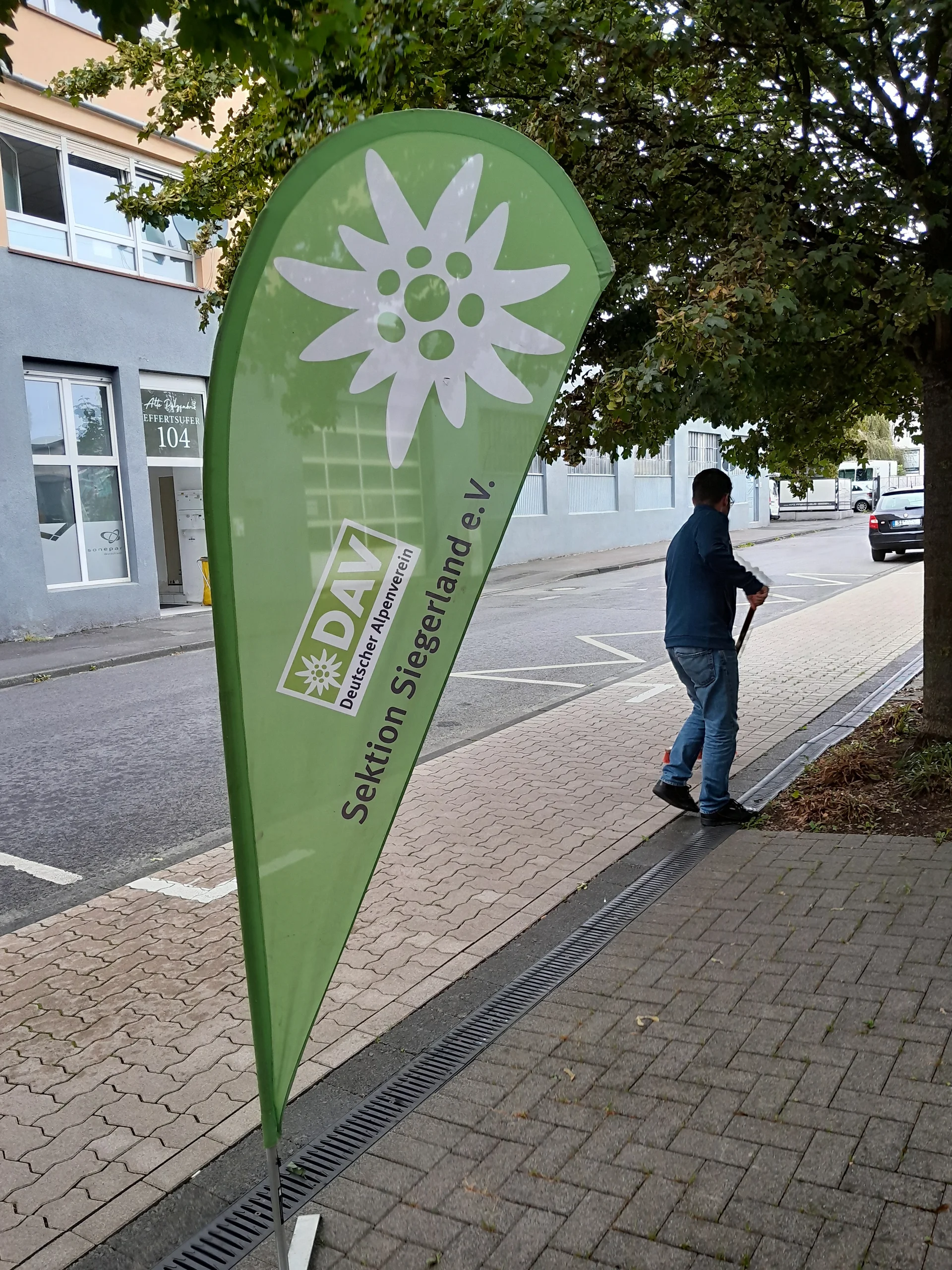 Ein grünes, tropfenförmiges Banner des Deutschen Alpenvereins (DAV), Sektion Siegerland e.V., steht am Straßenrand. Darauf ist in Weiß das Edelweiß-Logo des DAV abgebildet. Im Hintergrund ist ein Gebäude mit der Adresse Effertsufer 104 zu sehen. Rechts daneben fegt eine Person den Gehweg, teilweise von einem Baum überschattet. | © DAV Kletterzentrum Siegerland
